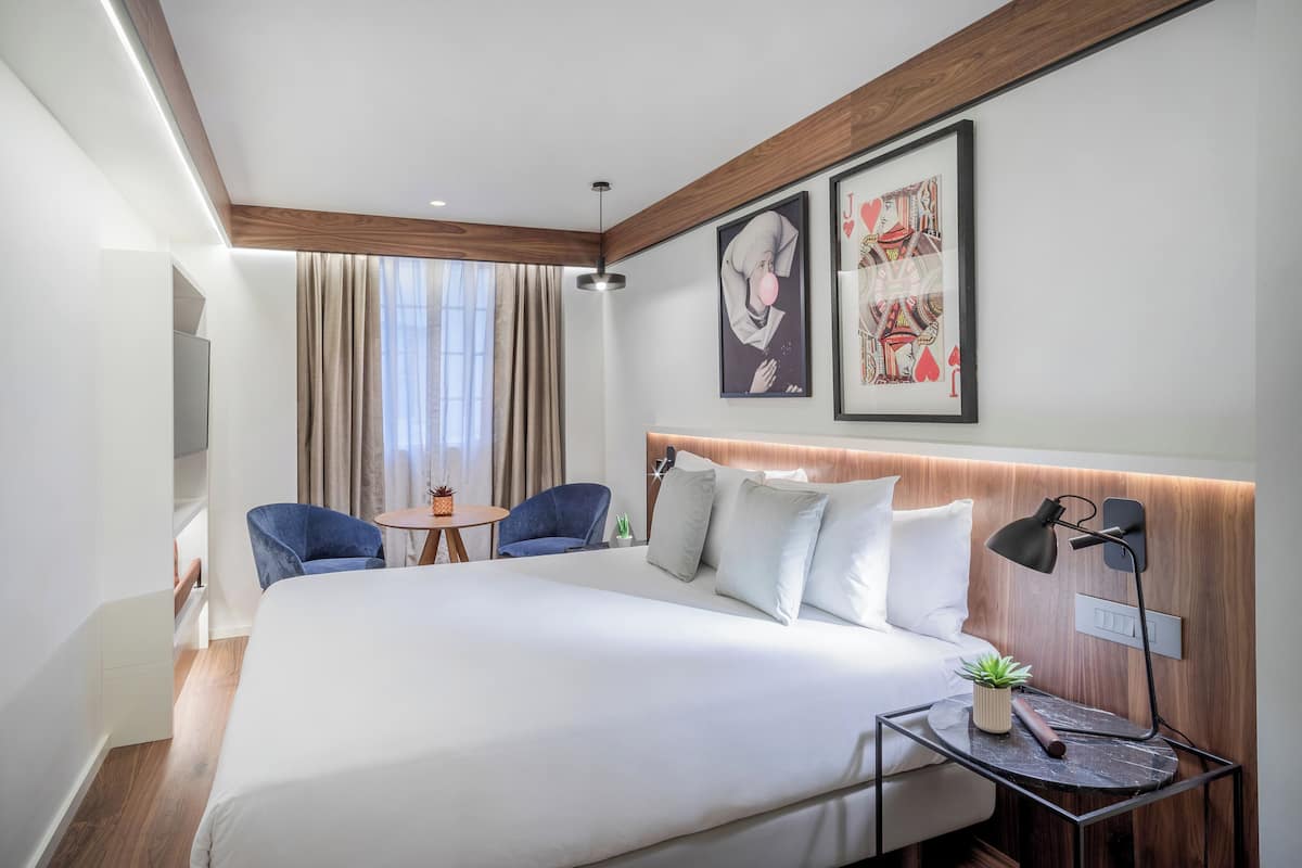 A modern hotel room with a large bed, a desk with a lamp, a chair, and a small table with a plant.