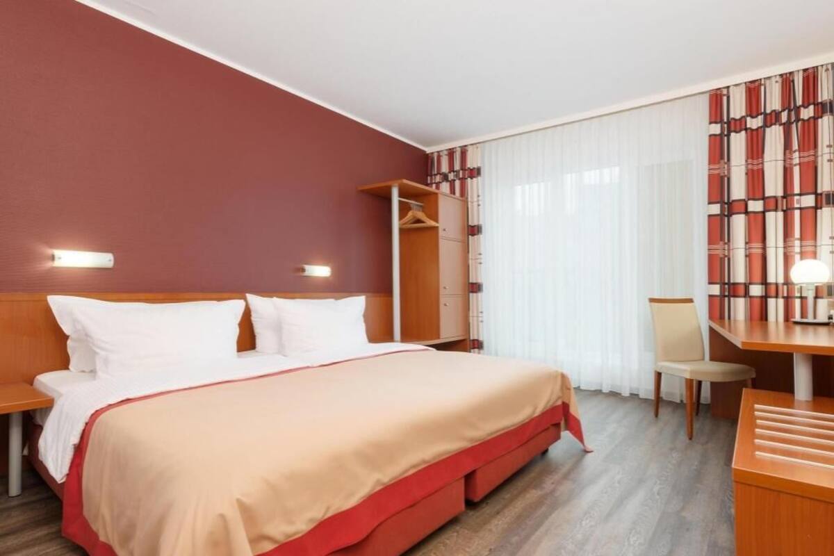 A hotel room with a bed, a desk, a chair, a wardrobe, and a window with curtains.