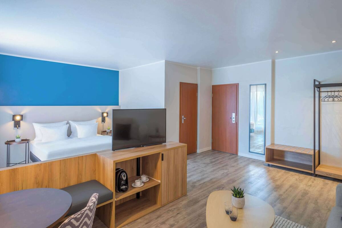 A modern hotel room with a bed, TV, wooden console, seating area, and a small table with a plant.