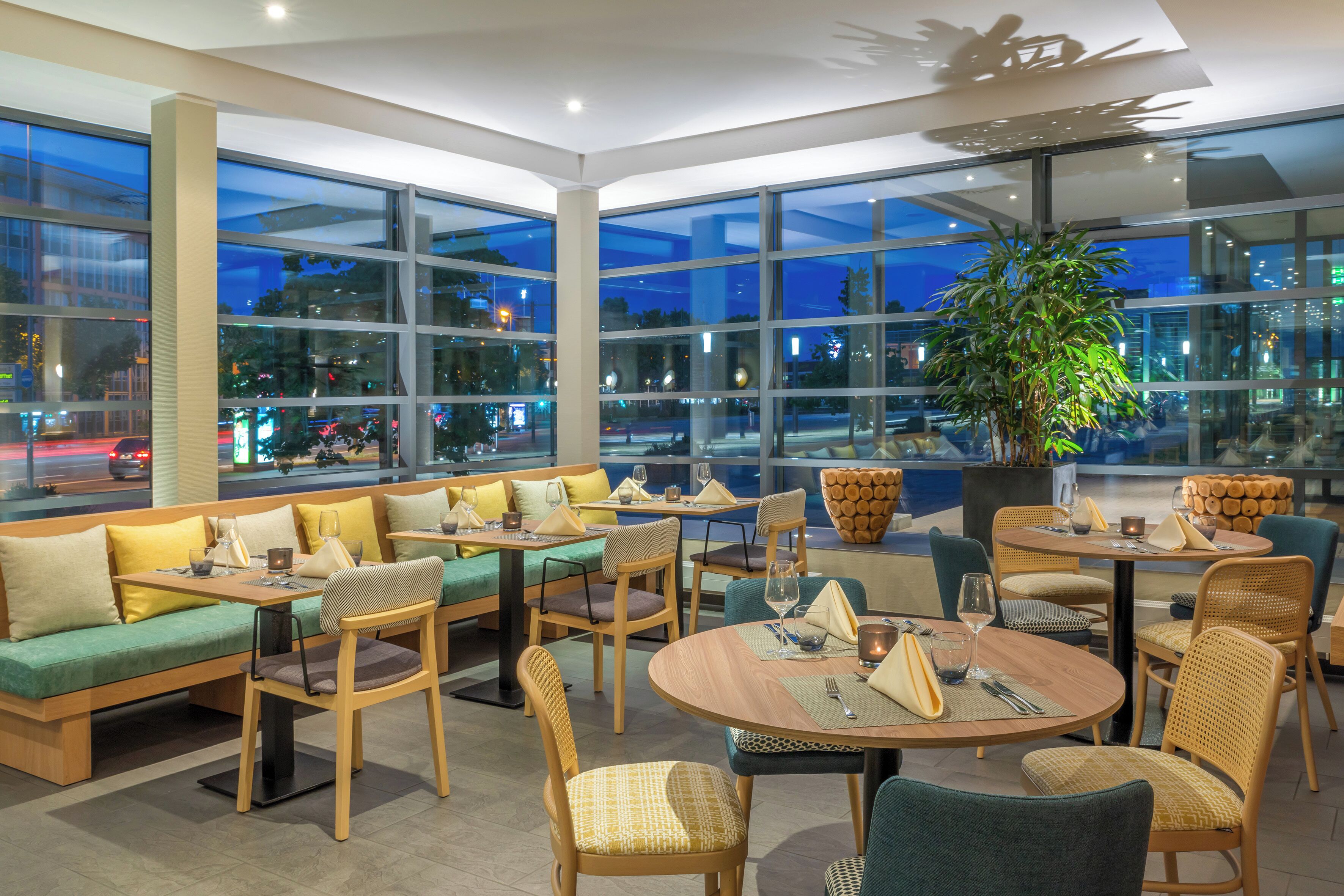 A modern indoor dining area with large windows, comfortable seating, and a large potted plant.