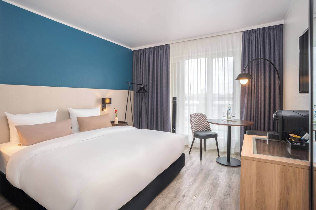 A hotel room with a large bed, a desk with a printer, a chair, and a table with a small table lamp.