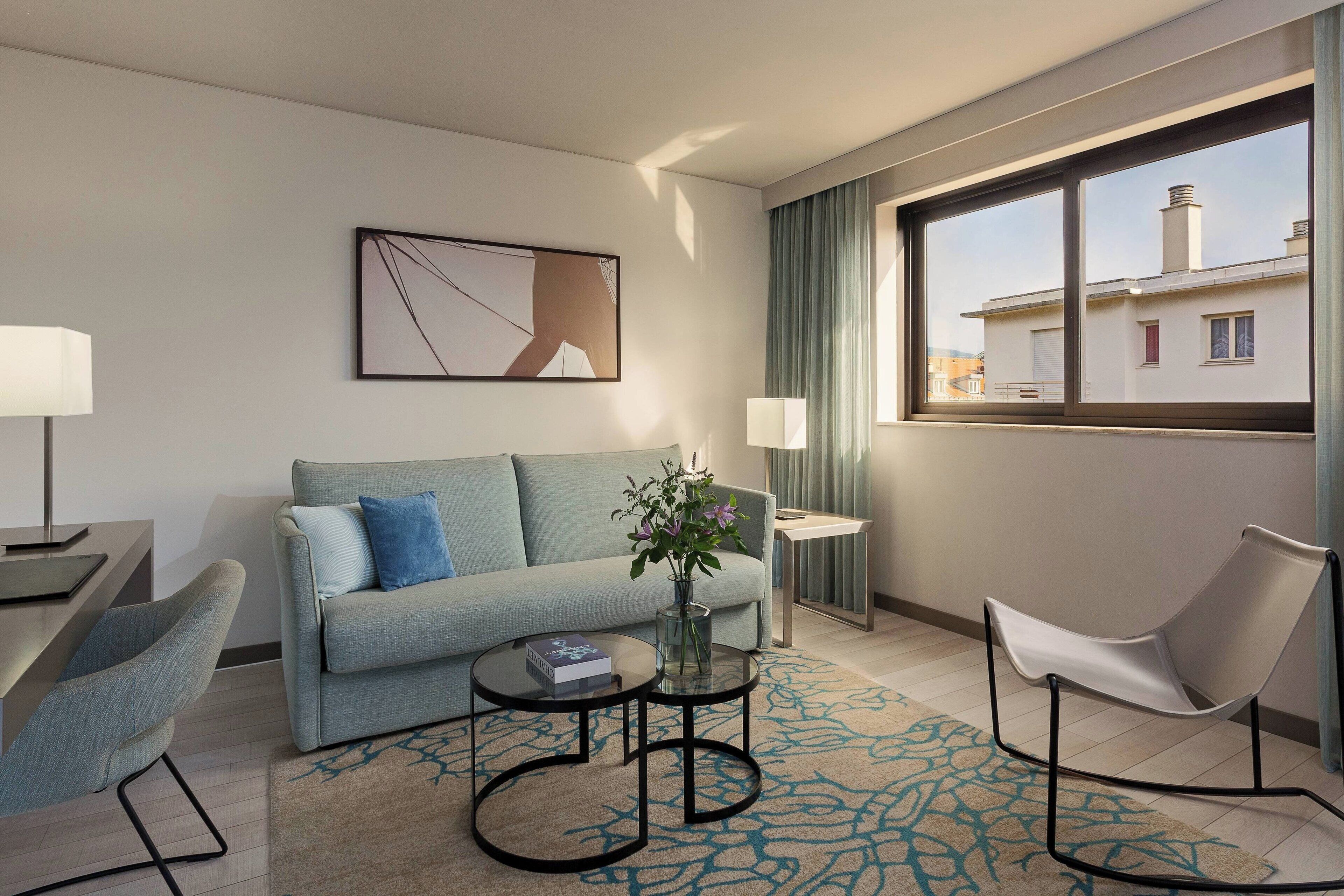 A modern hotel room with a sofa, a coffee table, a chair, and a large window offering a view of buildings.