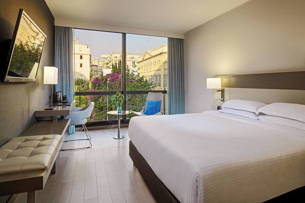 A hotel room with a large bed, a seating area with a chair and a small table, a TV mounted on the wall, and a view of the city through large windows.