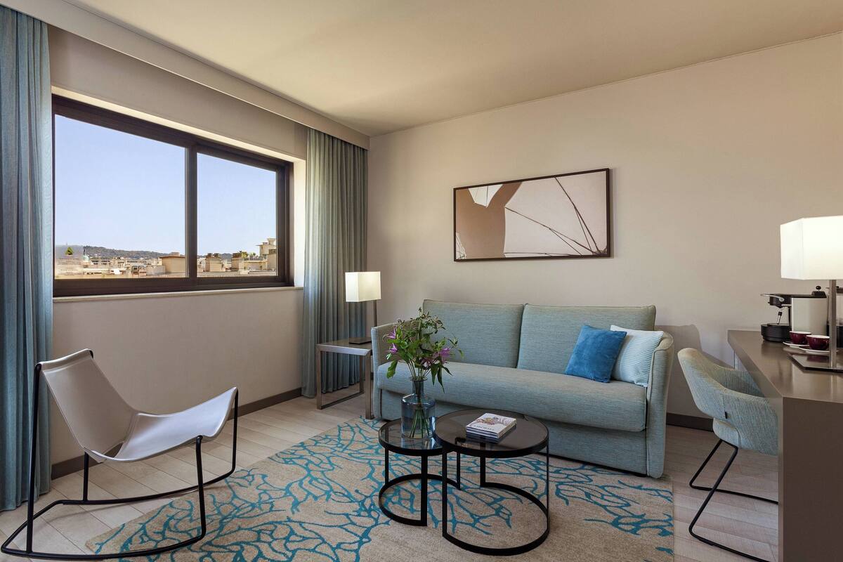 A modern hotel room with a sofa, a chair, a small table, a lamp, and a view of the cityscape.
