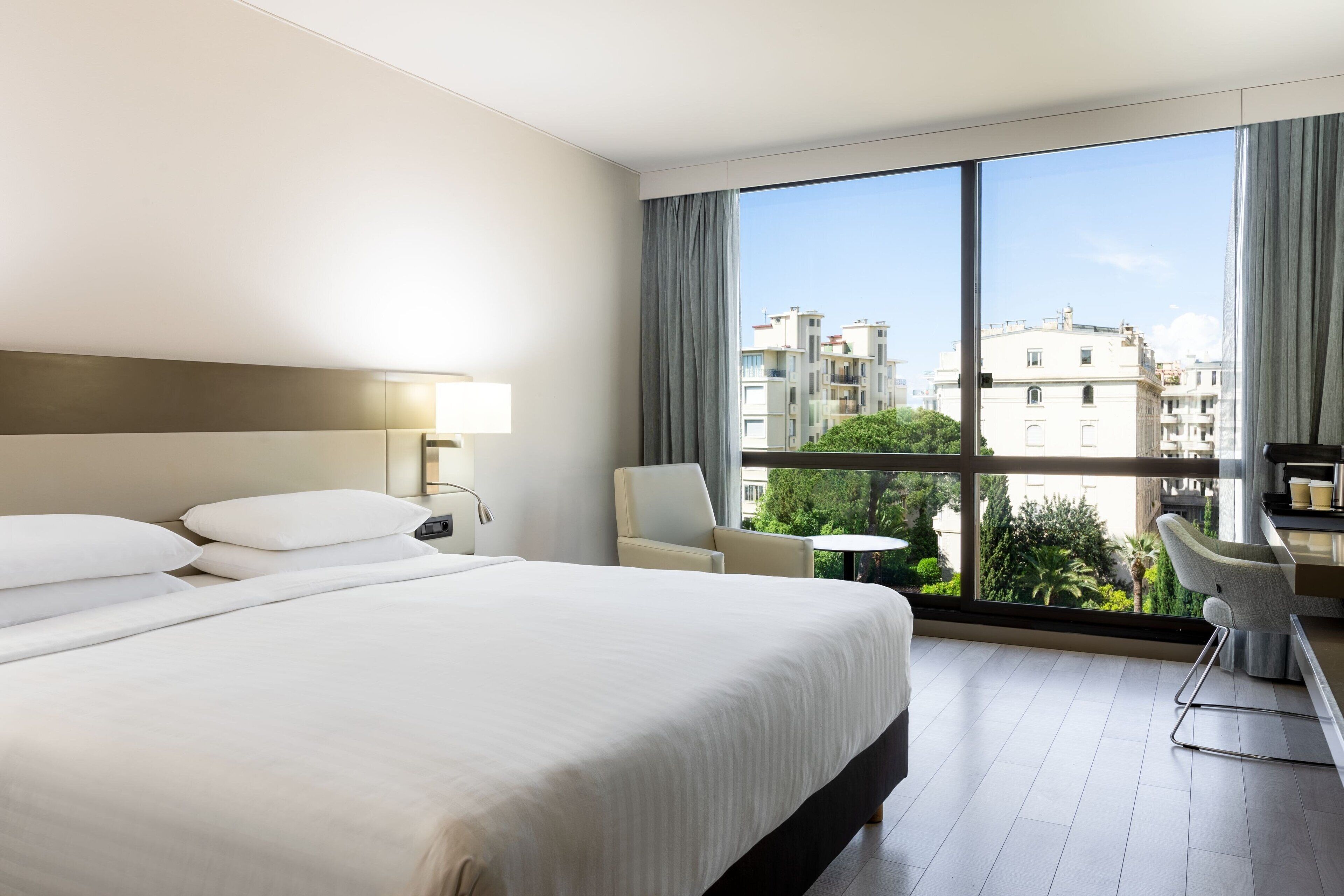 A hotel room with a large bed, a desk, a chair, and a view of the city through large windows.
