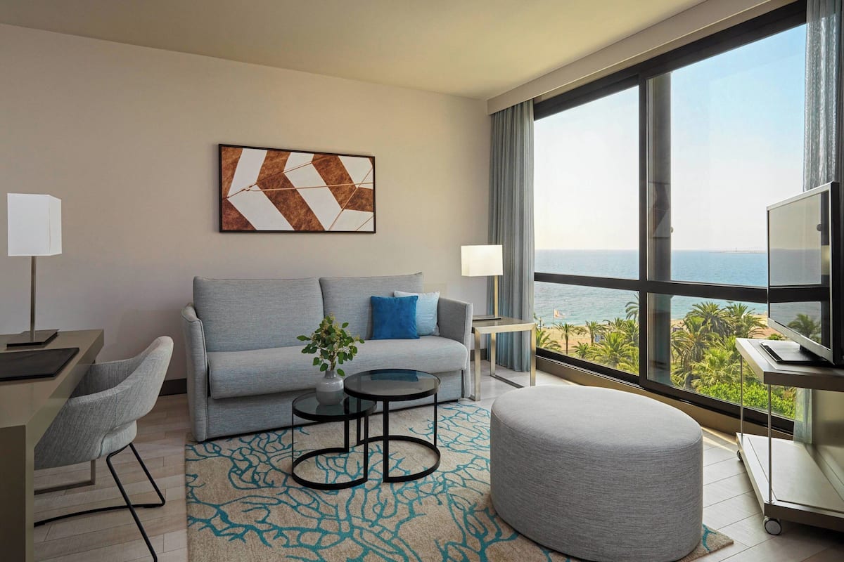 A modern hotel room with a grey sofa, a round ottoman, a small table with a plant, a TV on a cart, and a large window with a view of the sea.