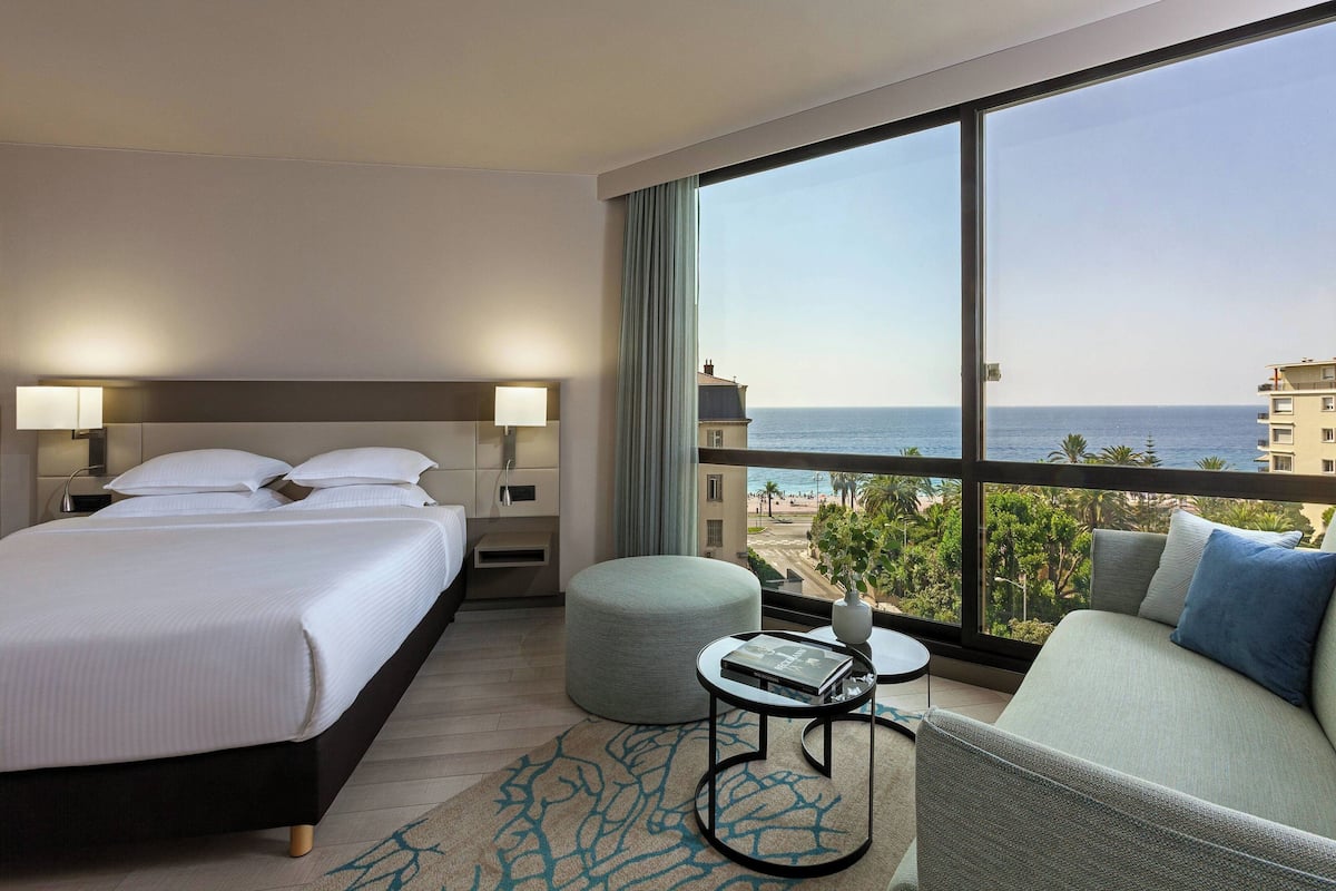 A hotel room with a bed, a sofa, a small table, and a large window offering a view of the sea and buildings.