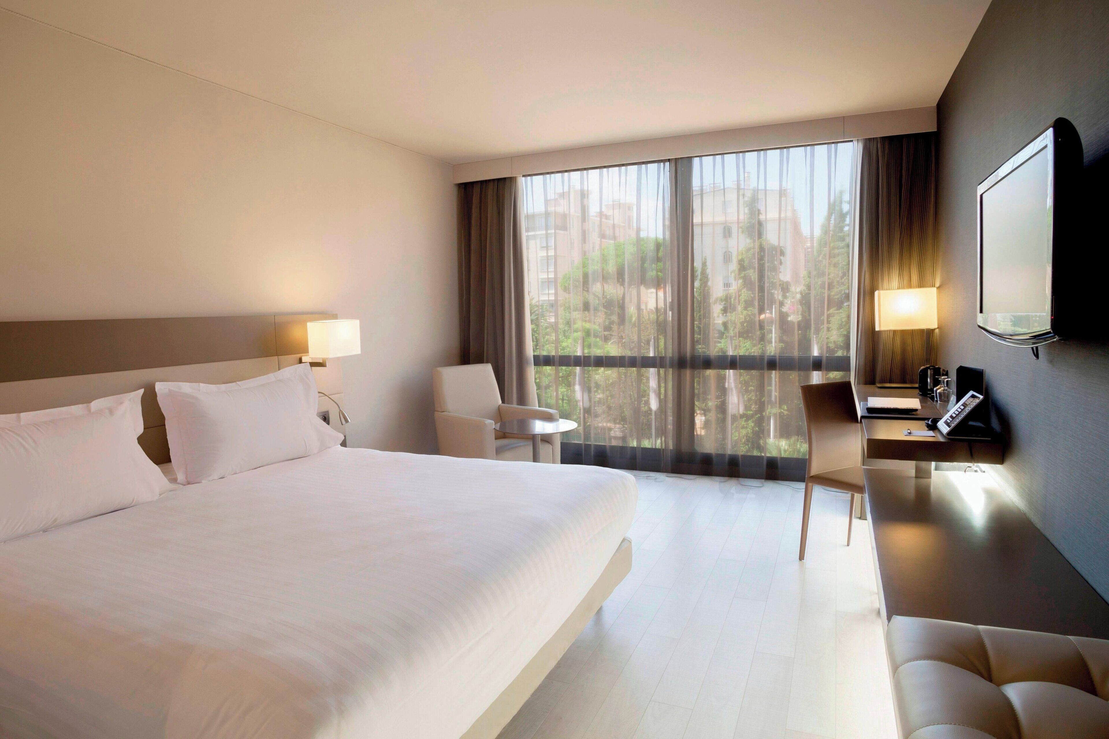 A modern hotel room with a large bed, a desk, a chair, and a TV mounted on the wall.