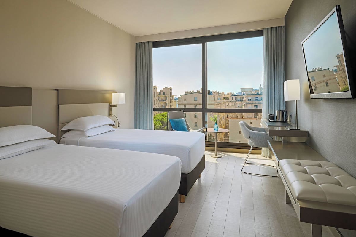 A hotel room with two beds, a sofa, a TV, and a balcony with a view of the city.