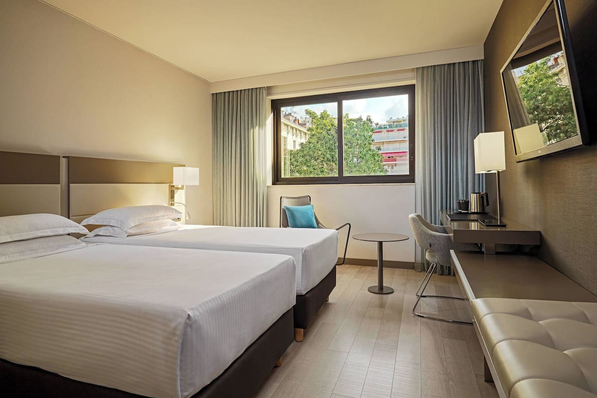 A hotel room with two beds, a desk, a chair, a small table, a TV, and a window with curtains.