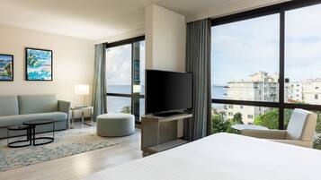 Una habitación de hotel moderna con una cama grande, un televisor sobre un mueble, un sofá, una pequeña mesa redonda y vistas al mar y a edificios.