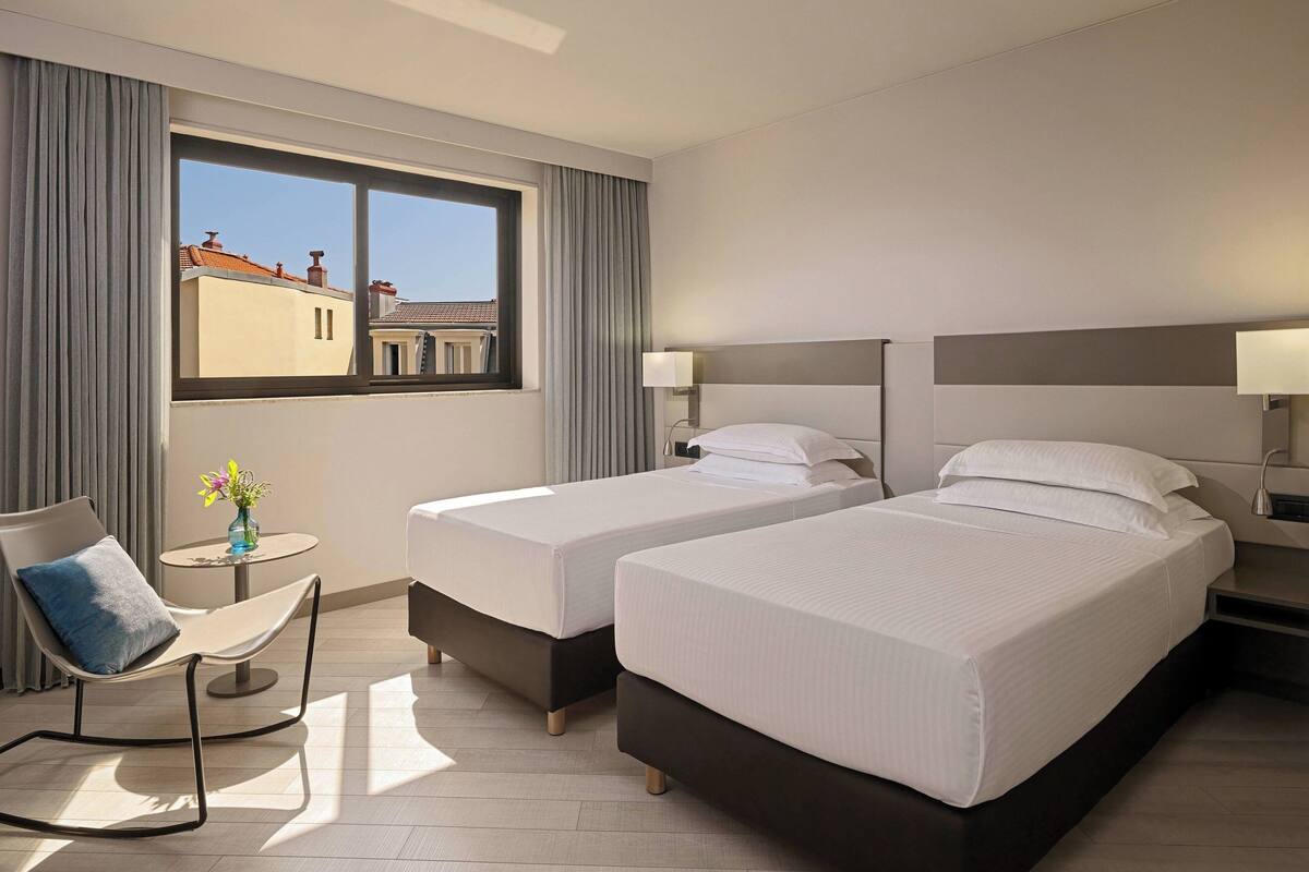 A hotel room with two beds, a chair, a small table with a vase, and a window with a view of buildings.