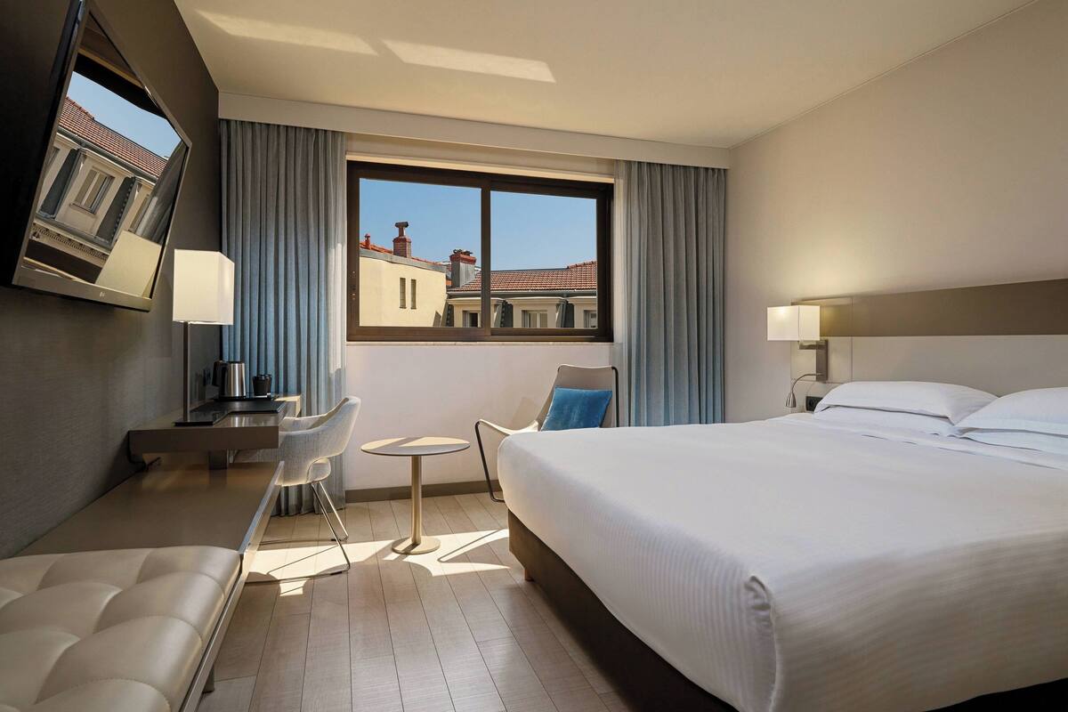 A hotel room with a bed, a desk, a chair, a small table, and a window with curtains.