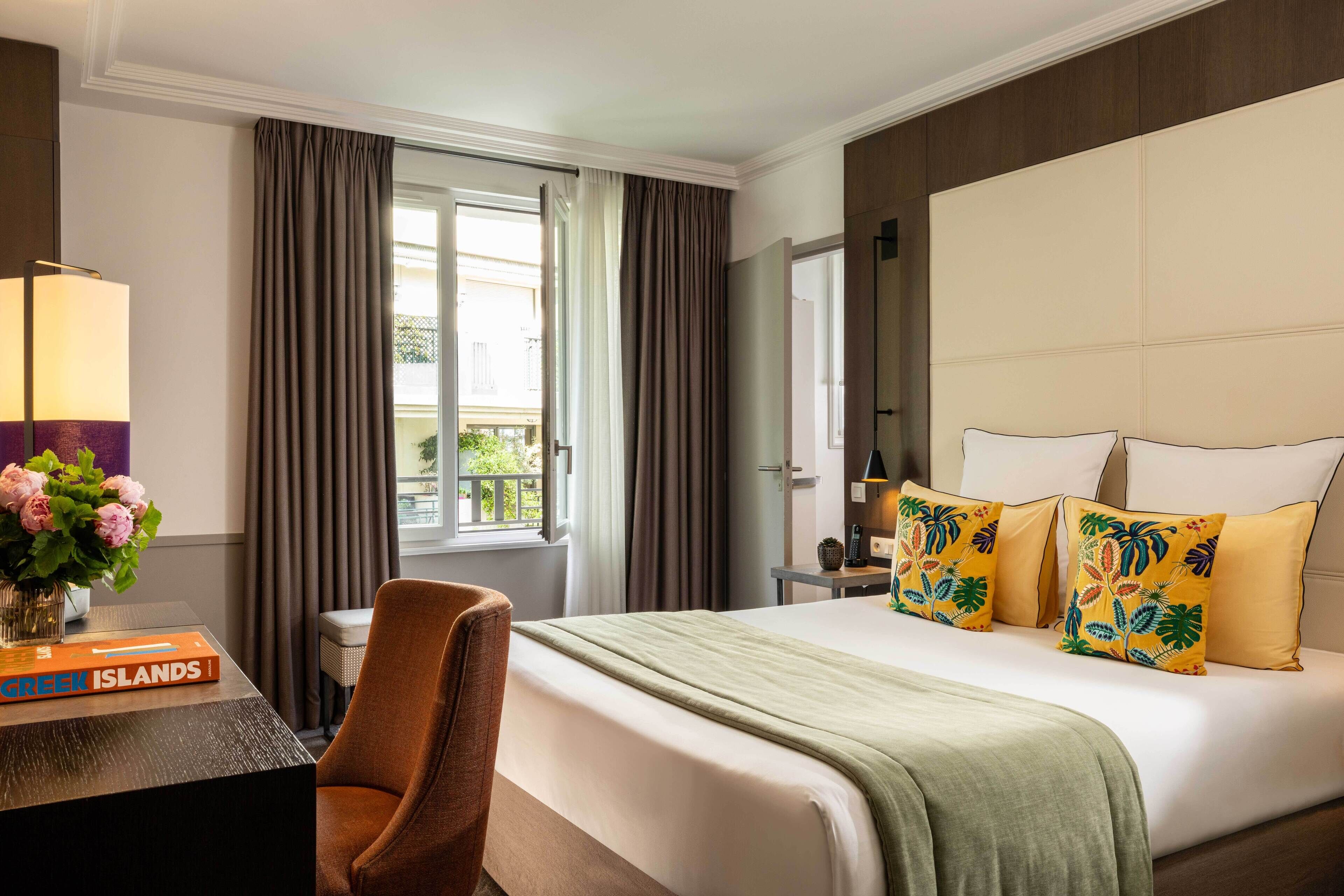 A hotel room with a large bed, a desk with a lamp, a chair, and a view of the outdoors through a window.