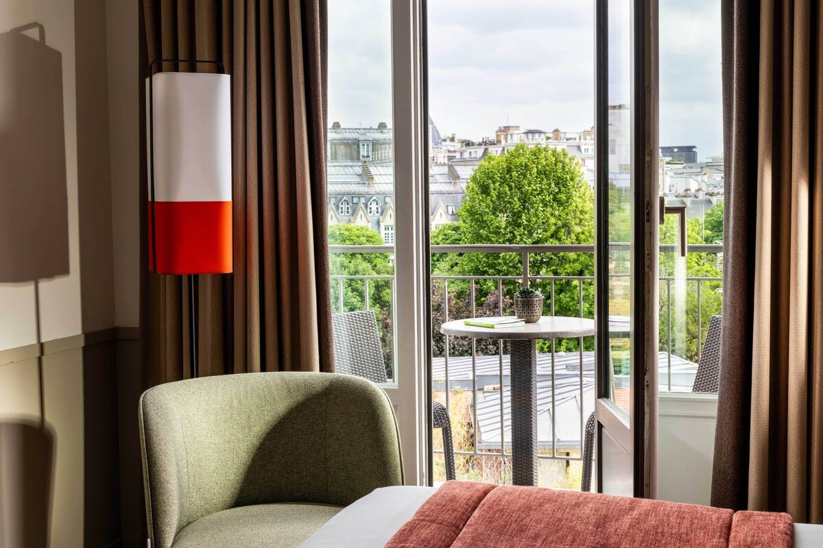 A hotel room with a large window, a green armchair, a small round table, and a view of a cityscape.