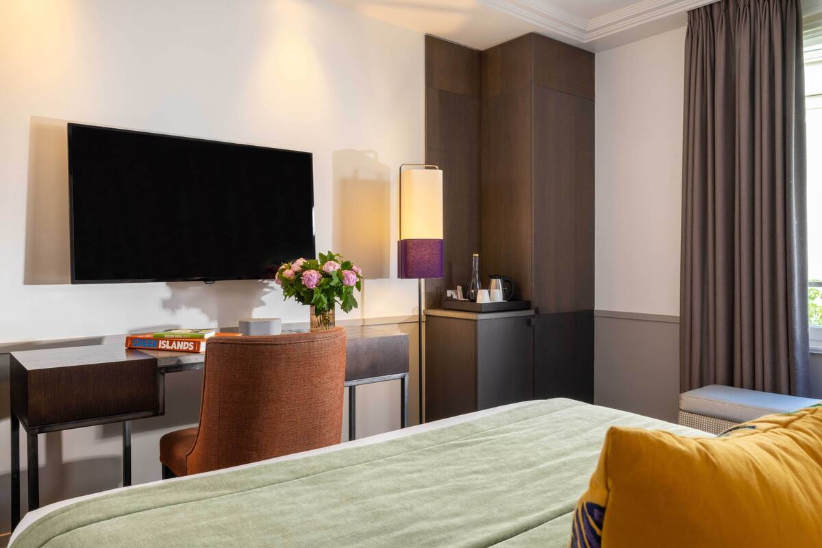 A hotel room with a bed, a desk, a chair, a TV, a lamp, a vase with flowers, and a window with curtains.