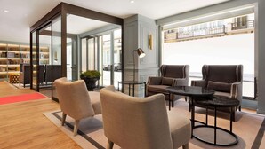 A modern interior with a seating area featuring armchairs and a round table, a bookshelf, and a view of the outside balcony. - Hotel Avenue Louise Brussels - Trademark Collection by Wyndham (Brussels)