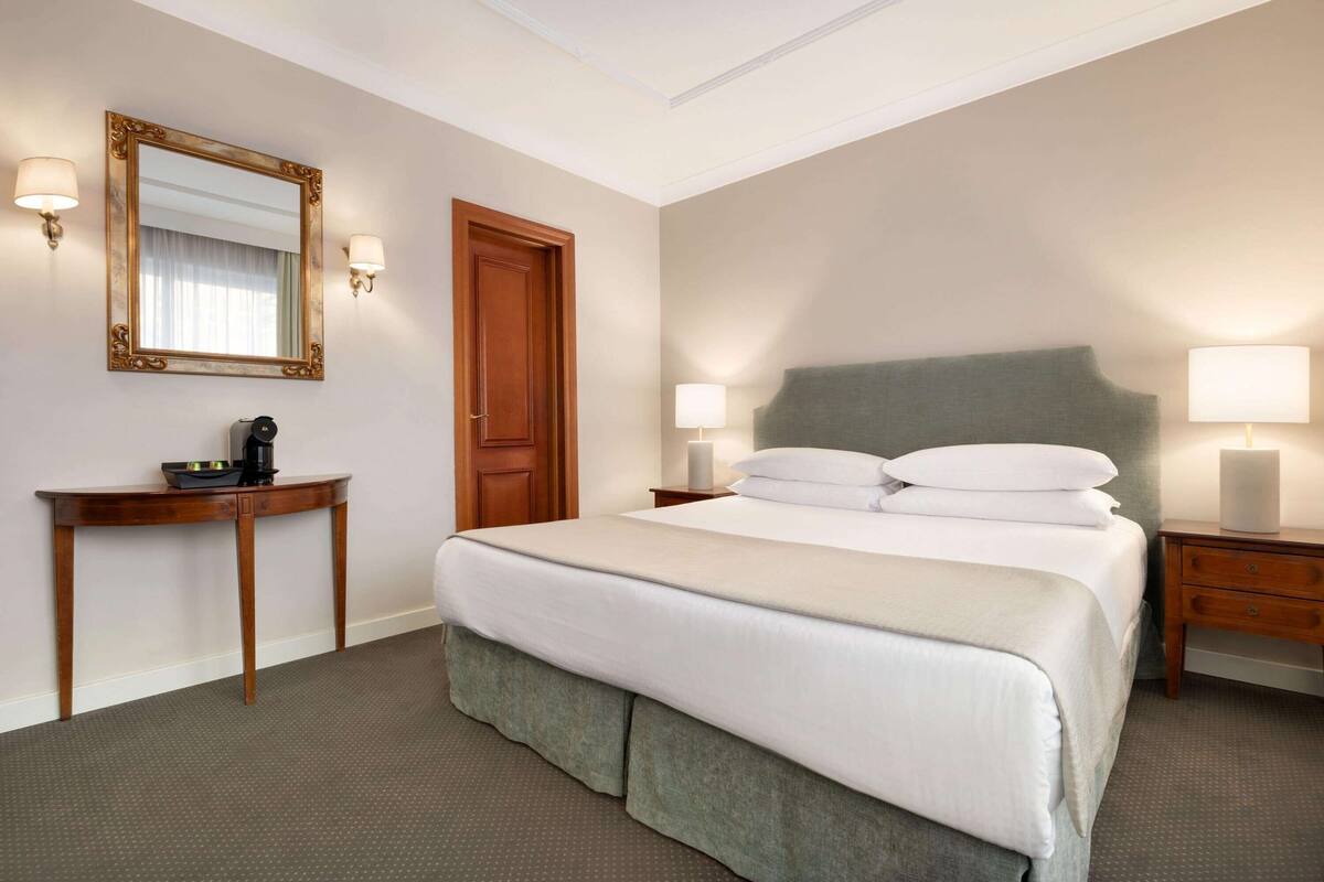 A hotel room with a large bed, bedside lamps, a wooden nightstand, a small table with a coffee maker, and a mirror on the wall.