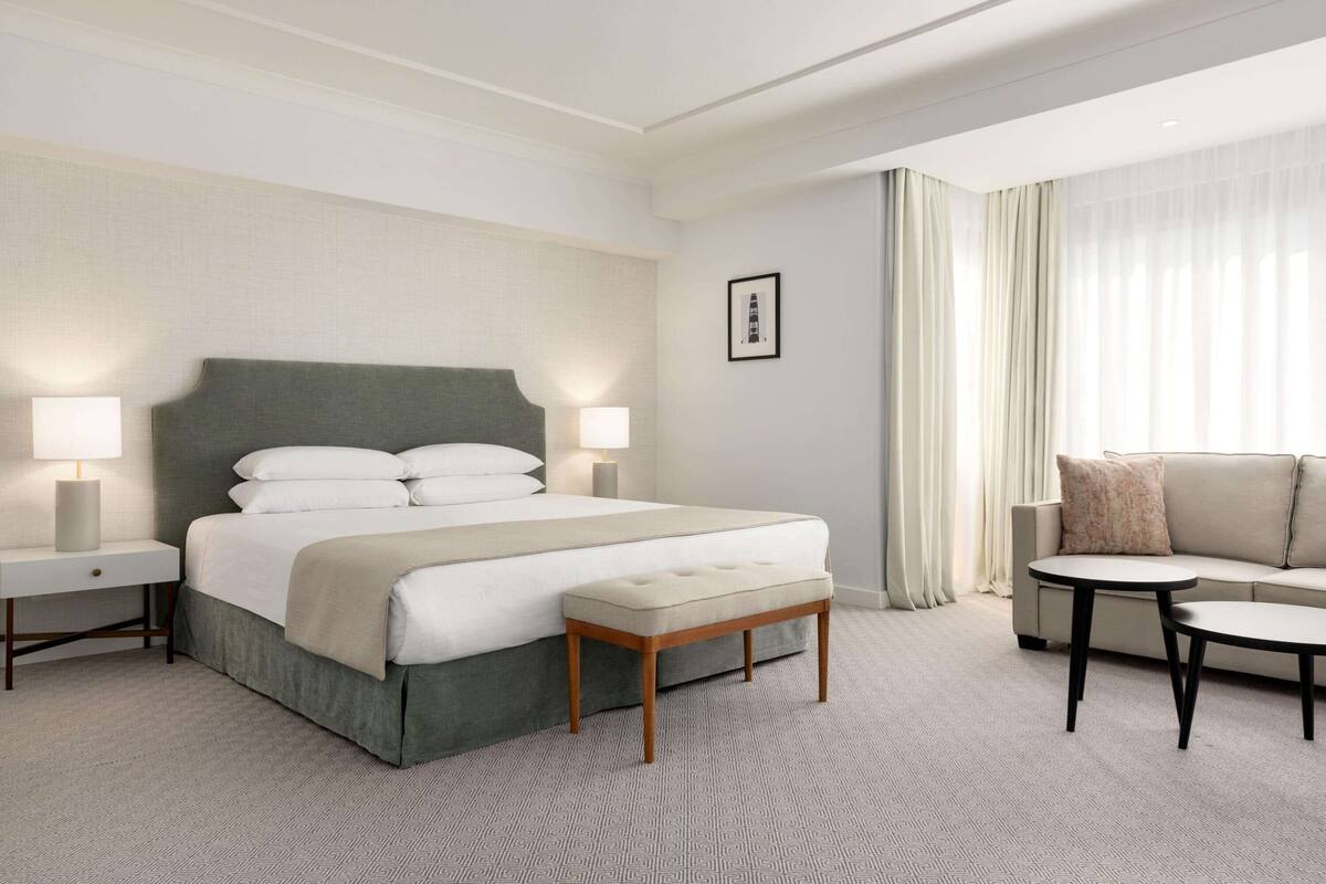 A hotel room with a large bed, bedside tables, a bench, a sofa, and a small table.