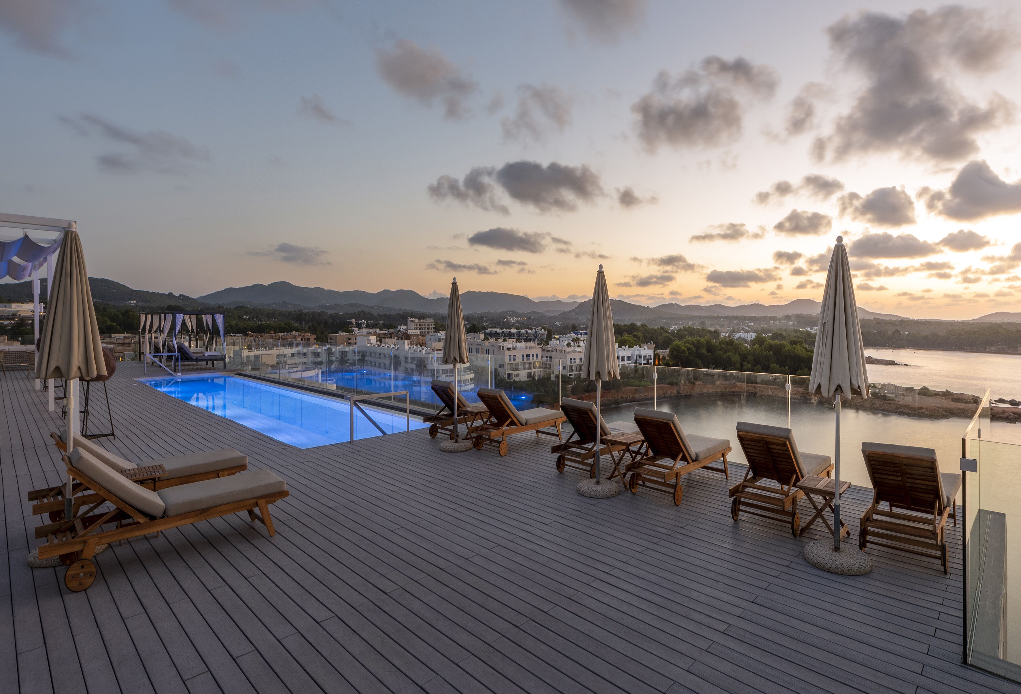 A rooftop pool area with lounge chairs and umbrellas, overlooking a body of water and distant mountains.