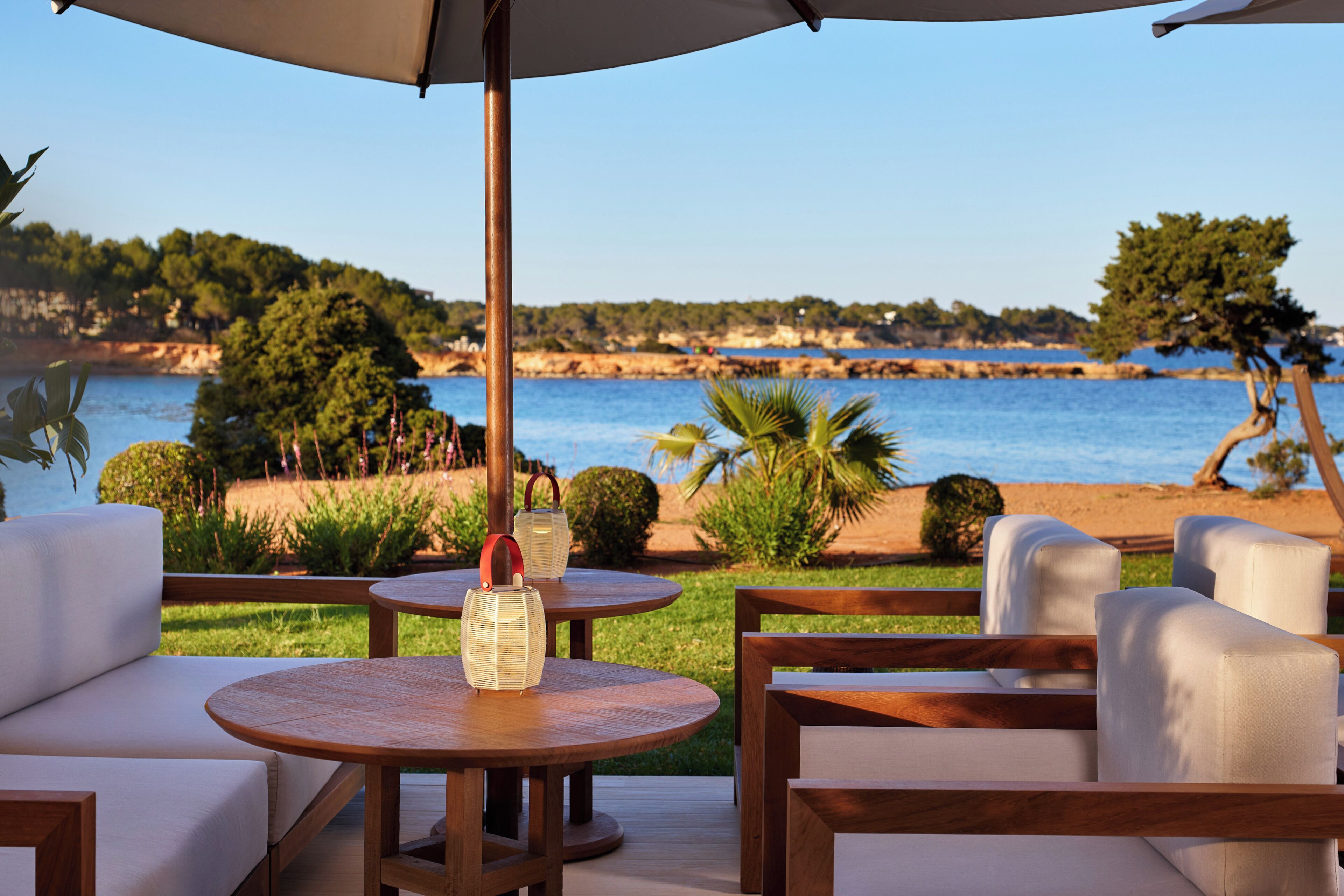 A seaside patio with round wooden tables, white cushioned seating, and a view of the sea and greenery.