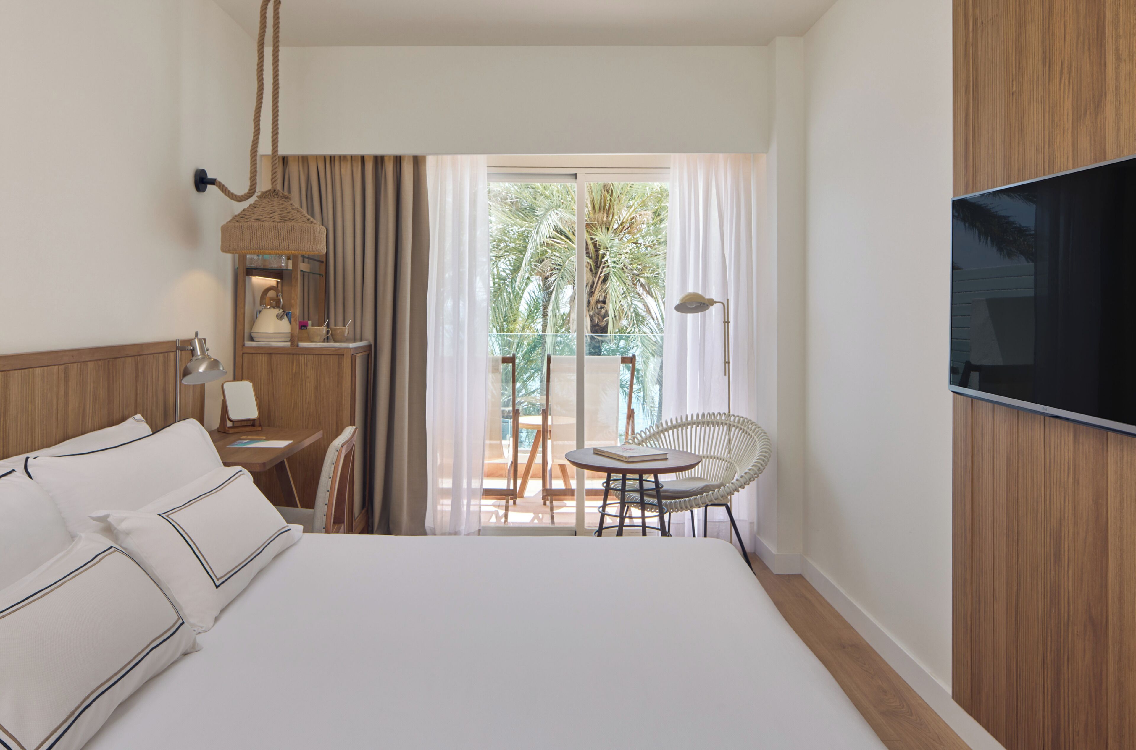 A hotel room with a large bed, a desk with a lamp, a chair, a TV, and a balcony with a chair and table.