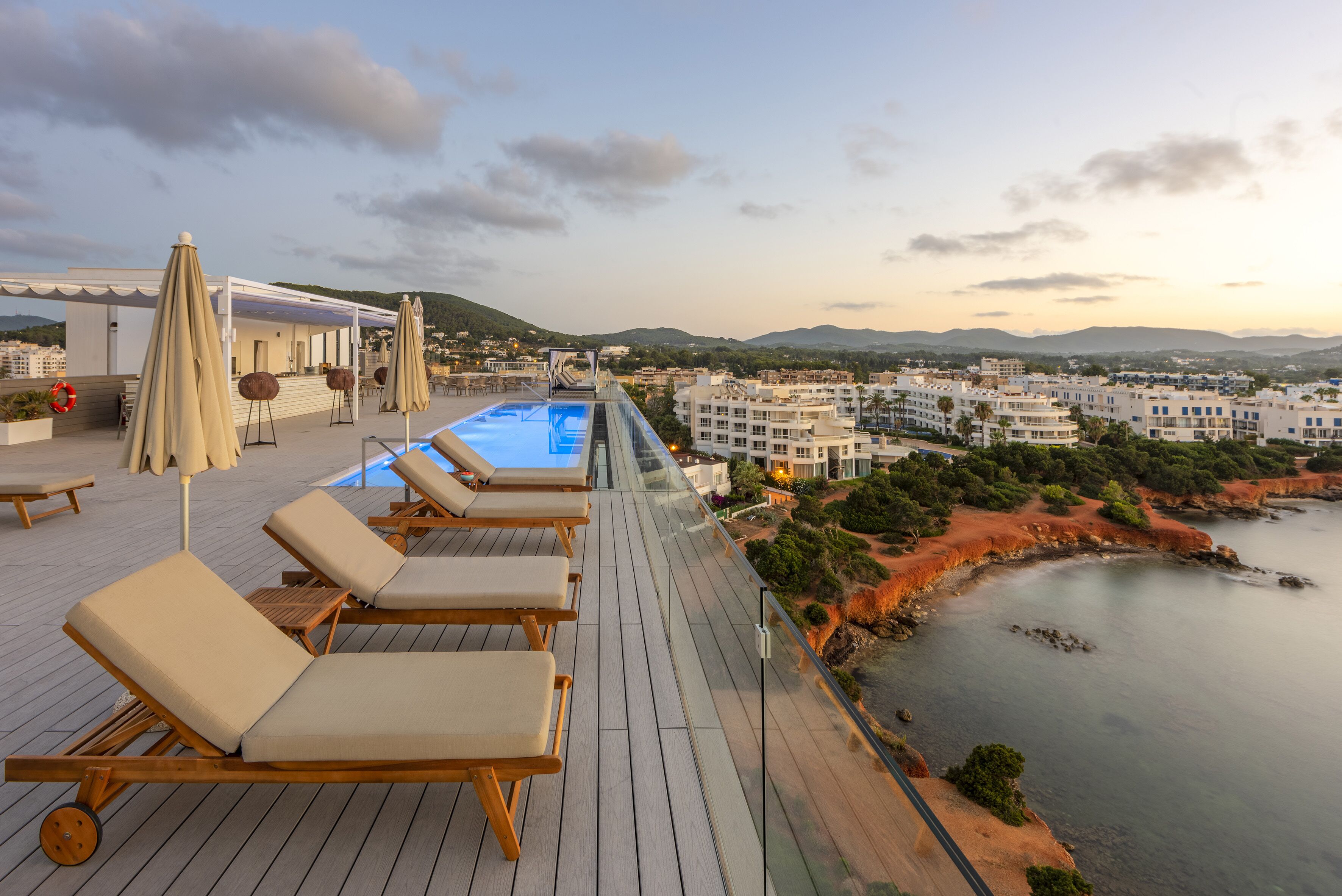 A rooftop pool area with lounge chairs and umbrellas, overlooking a coastal town and hills.