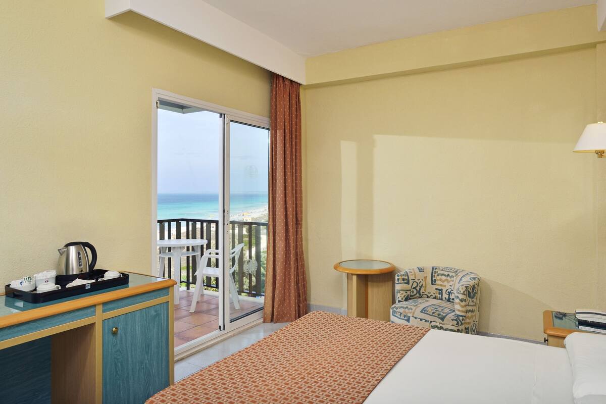 Ein Hotelzimmer mit Bett, Schreibtisch mit Wasserkocher, Stuhl, kleinem Tisch und Blick auf den Strand.
