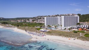 A coastal hotel with multiple floors, a beach in the foreground, and a hillside with residential buildings in the background. - Sol Milanos Pingüinos (Alayor)