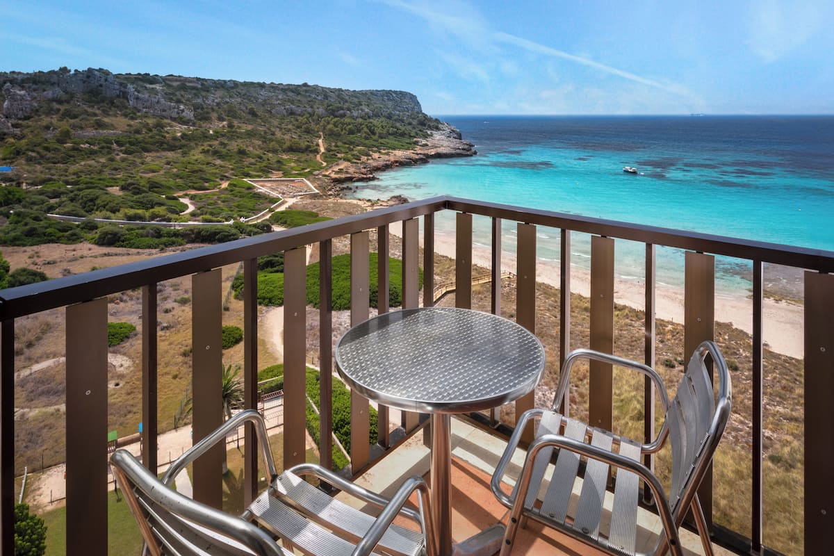 Ein Balkon mit einem Metalltisch und -stühlen, der auf eine Küstenlandschaft mit Strand und Klippen blickt.