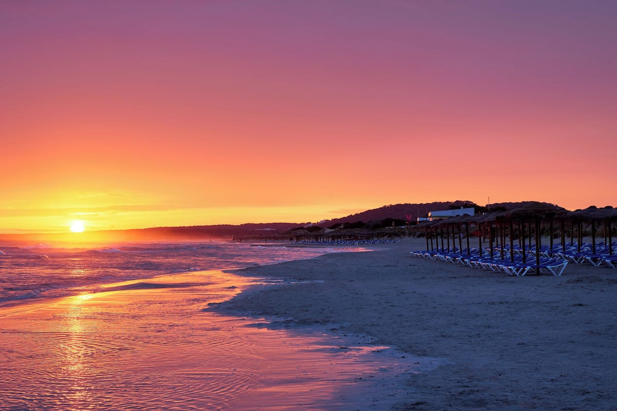 Ein Strand bei Sonnenuntergang mit einer Reihe von Liegestühlen und einem Gebäude im Hintergrund.