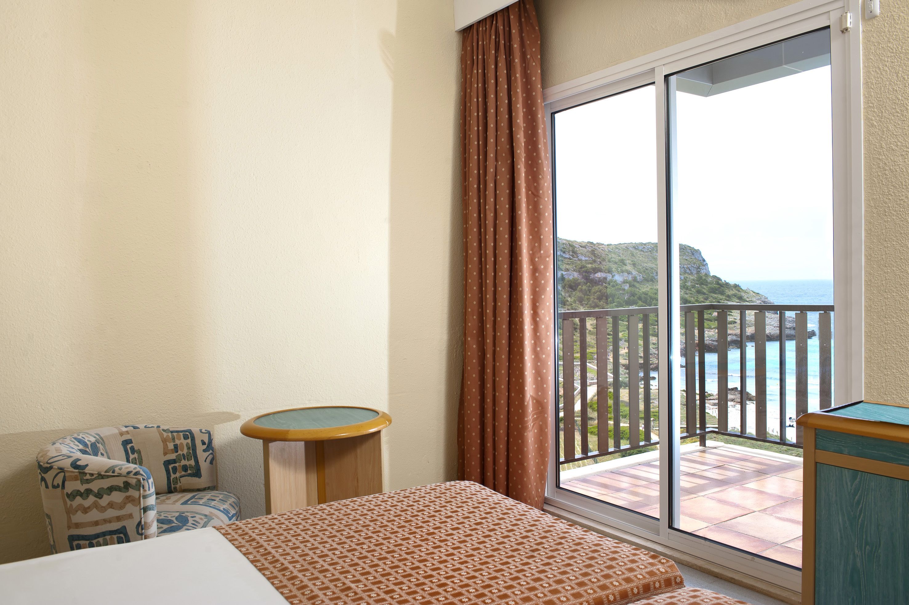A hotel room with a bed, a small table, a chair, and a balcony with a view of the sea and hills.