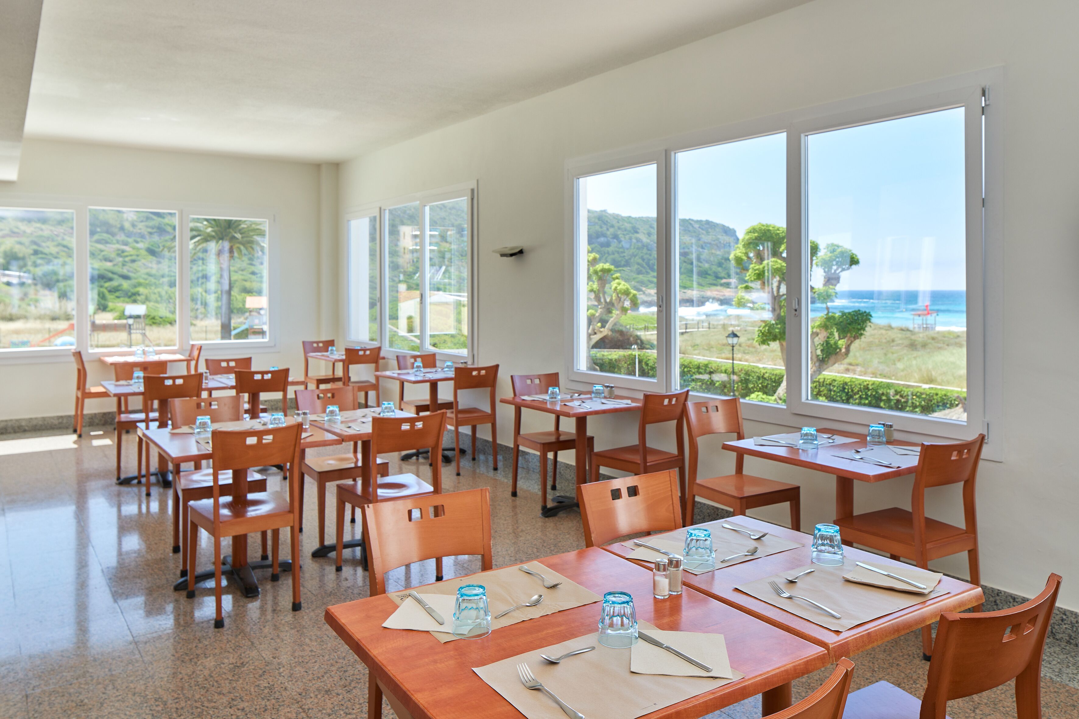 Un comedor con mesas y sillas, ventanas amplias con vistas a la playa y las colinas.