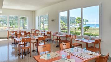 Un comedor con mesas y sillas, ventanas amplias con vistas a la playa y las colinas.