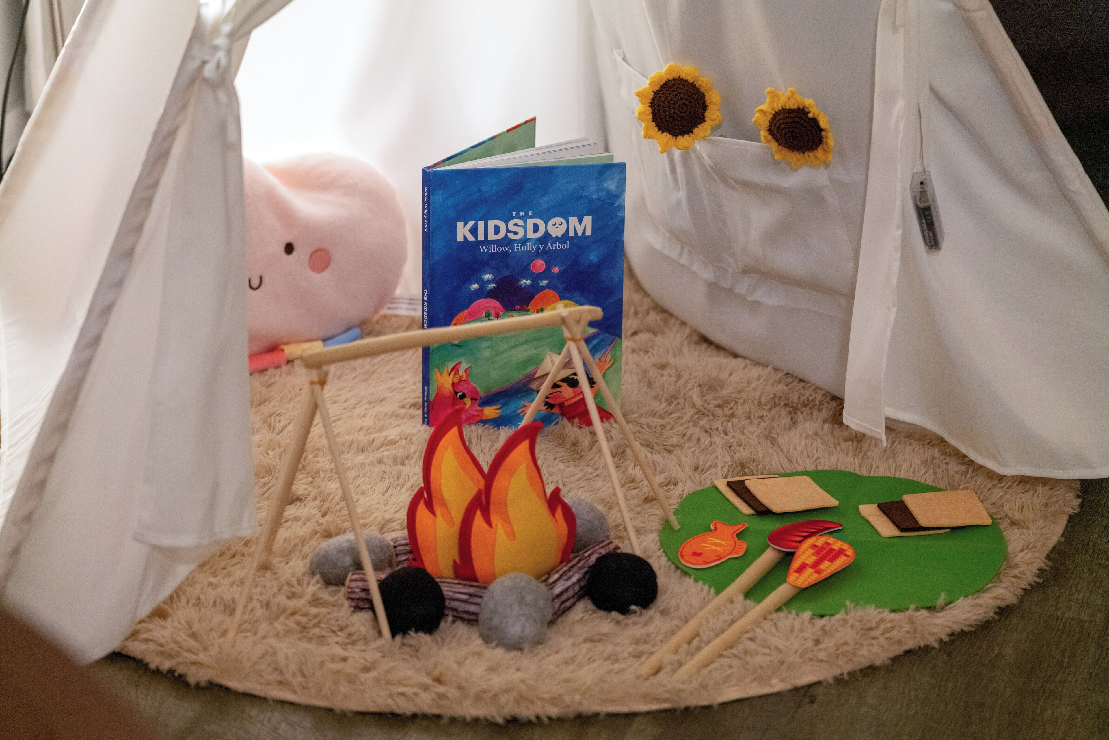 Un acogedor campamento dentro de casa con una tienda de campaña, un libro titulado 'The Kidsom' y una pequeña fogata.