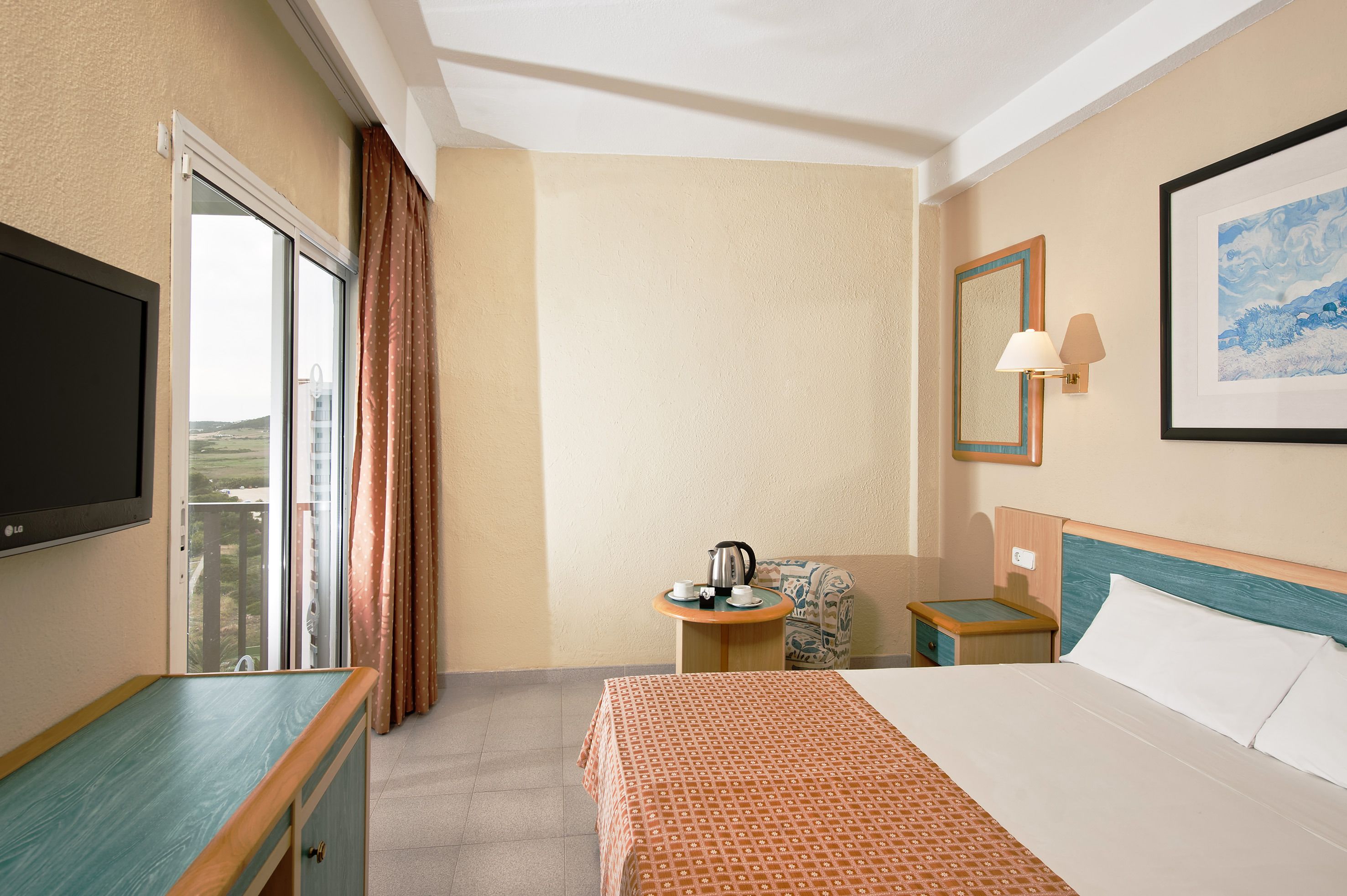 Une chambre d’hôtel avec un lit, une télévision, une table avec une bouilloire, une chaise et une fenêtre avec des rideaux.