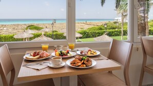 A breakfast table with plates of food, a cup of coffee, and a glass of orange juice, overlooking a beach with palm trees and a clear sky. - Sol Milanos Pingüinos (Alayor)