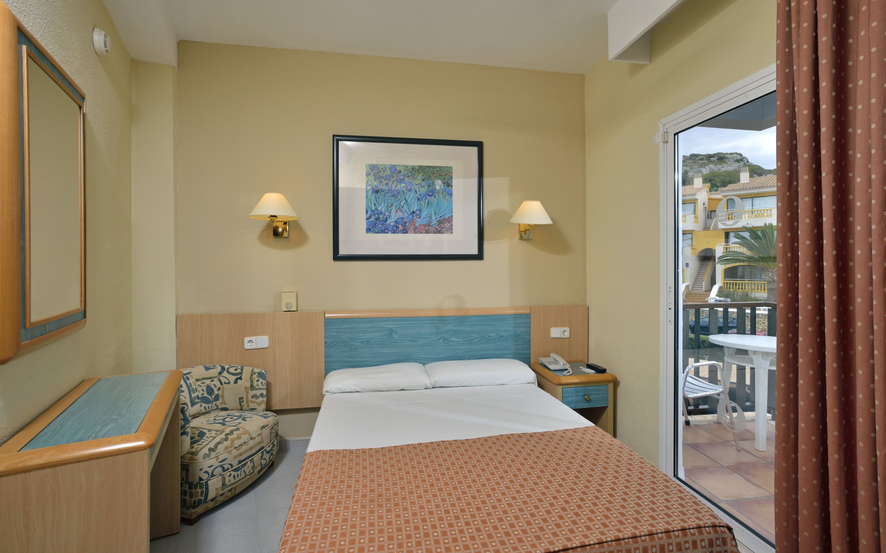 A hotel room with a bed, two bedside lamps, a framed artwork, a chair, and a view of the outside balcony.