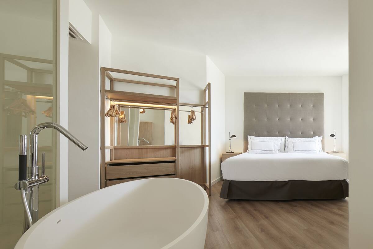 Una habitación de hotel moderna con una bañera grande, un espejo amplio, un cabecero capitonado y una cama bien tendida.