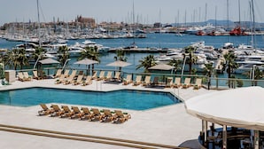 Un puerto deportivo con numerosas embarcaciones, una piscina con tumbonas y vistas a un paisaje urbano al fondo.