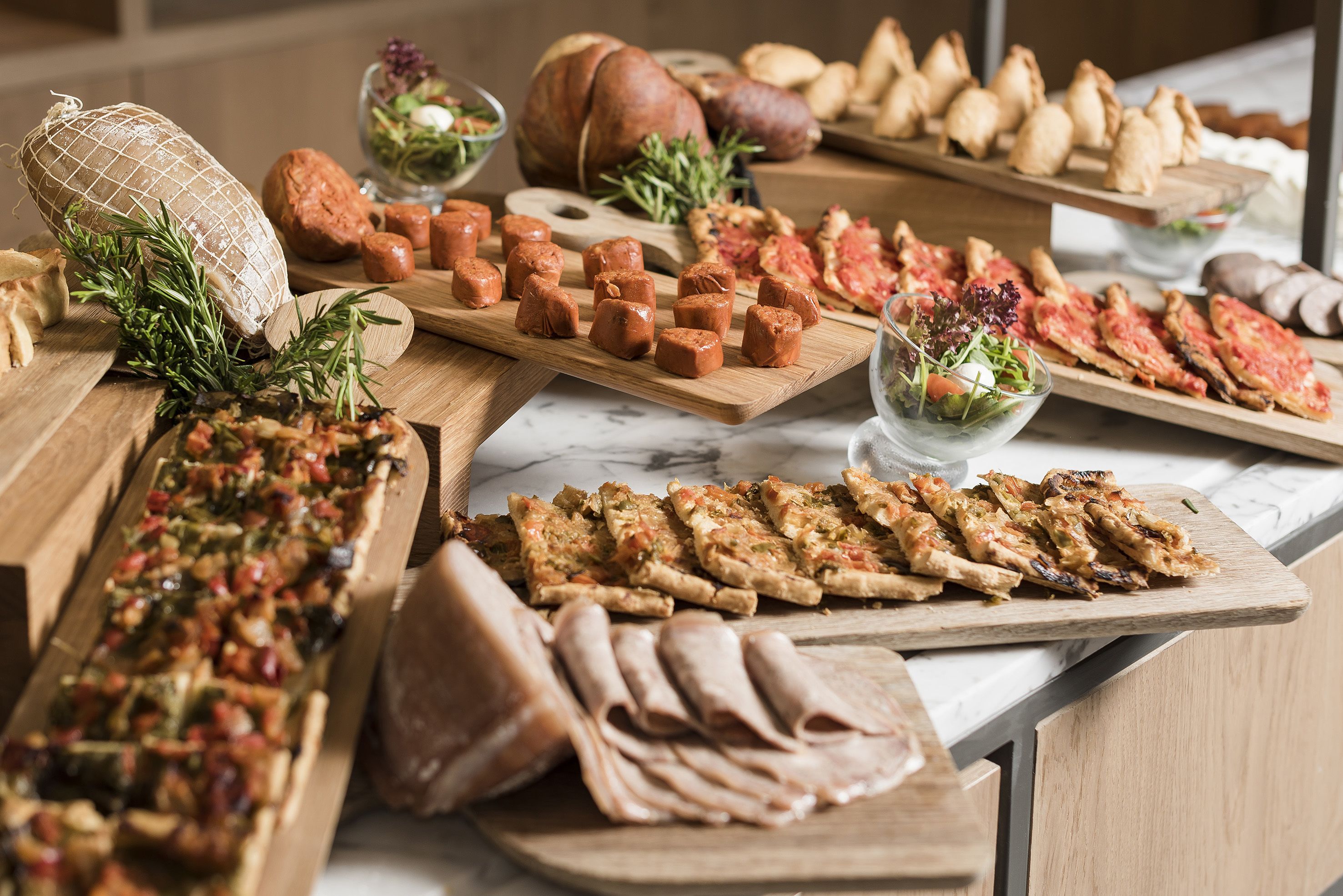Verschiedene Speisen auf Holztischen präsentiert, darunter Aufschnitt, Gebäck und ein Gemüsegericht.