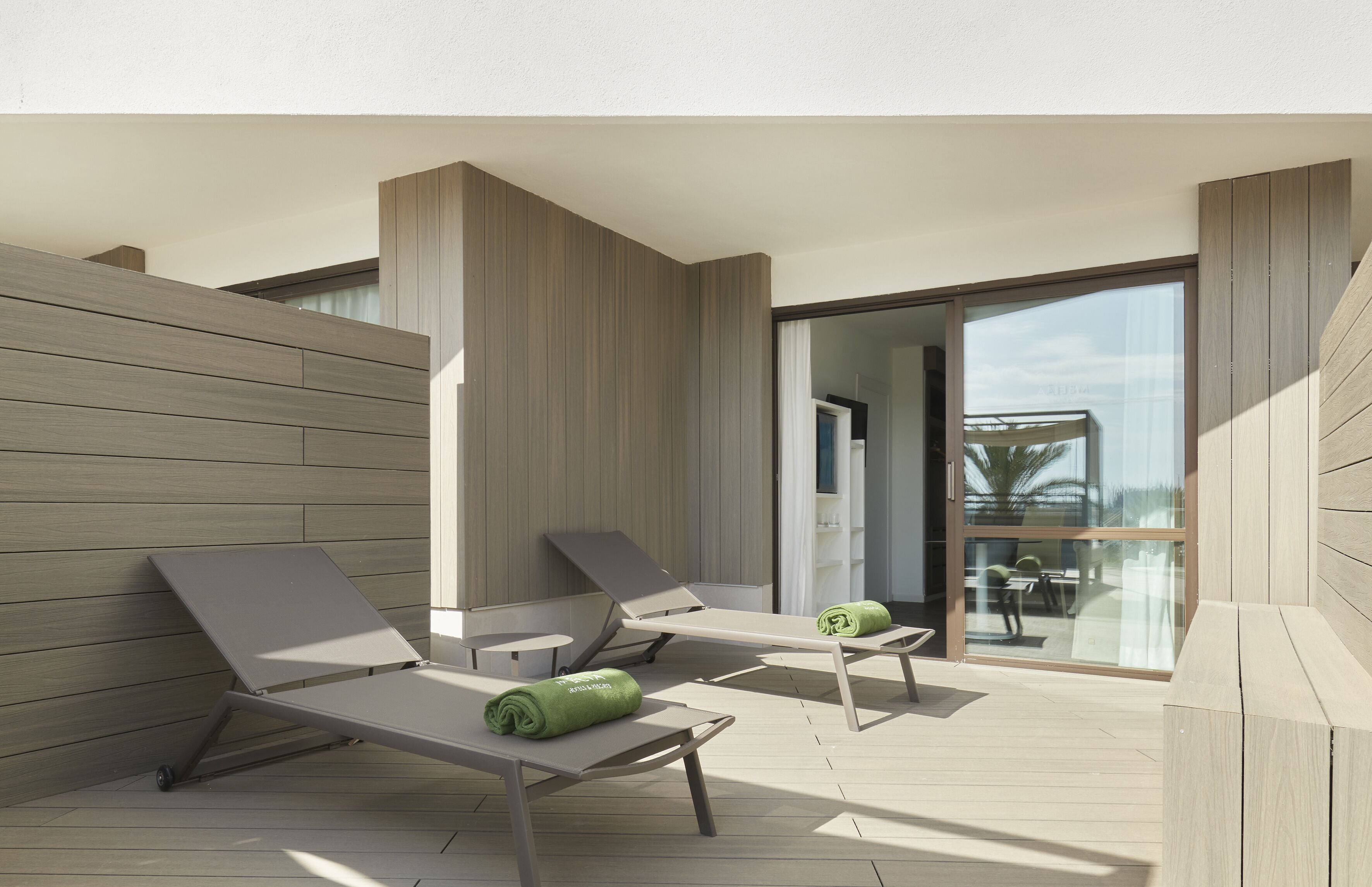 Un espacio lounge moderno al aire libre con dos tumbonas y una puerta de cristal que da a un ambiente interior.