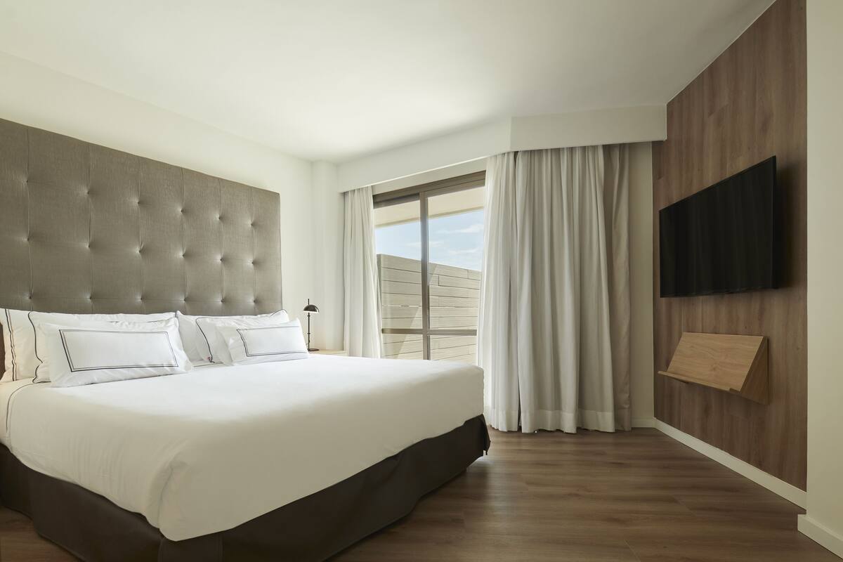 Habitación de hotel moderna con una cama grande, un televisor de pantalla plana montado en la pared y una puerta corrediza de vidrio que da a un balcón.