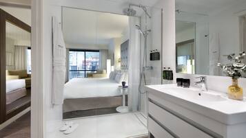 Una habitación de hotel moderna con ducha de vidrio, una cama grande, un tocador blanco con espejo y vistas de la ciudad a través de la ventana.