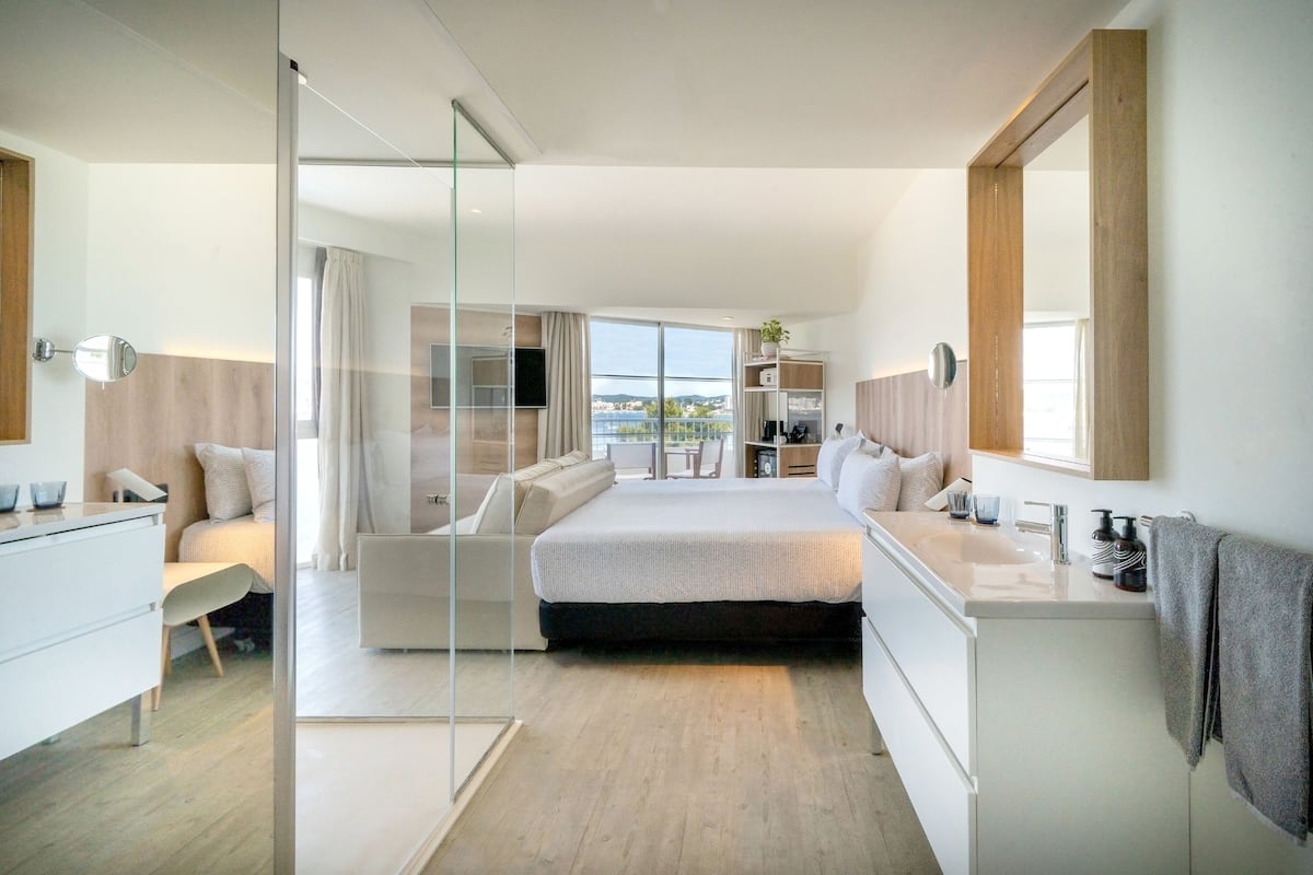 A modern bedroom with a large bed, a glass partition, a bedside table, a built-in TV, and a view of the outdoors.