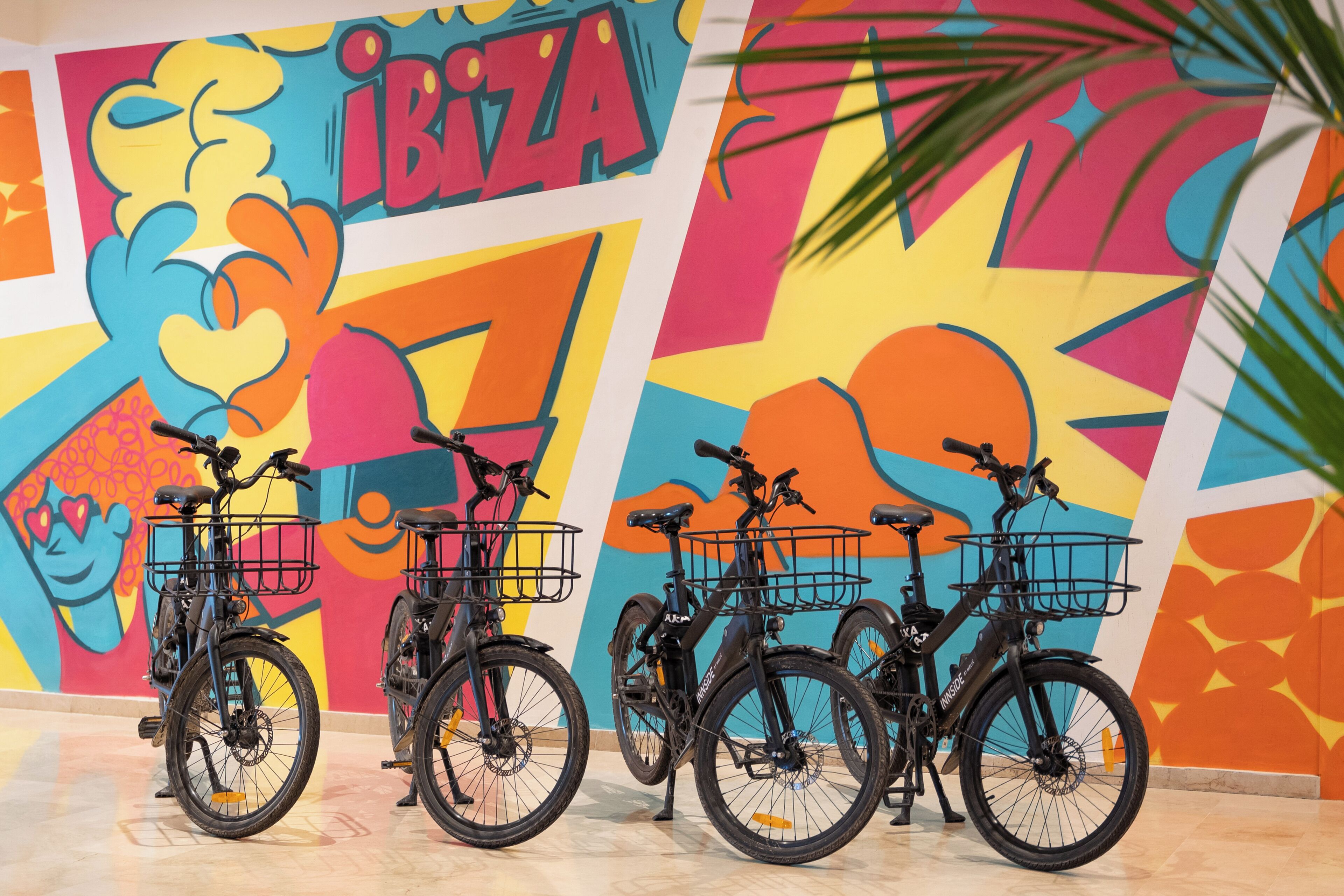 Four bicycles with baskets, parked against a colorful mural with the word "IBIZA" prominently displayed.
