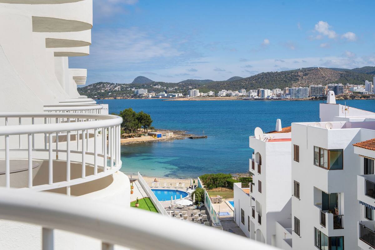 A coastal hotel with a pool, balconies, and a view of the sea and cityscape.