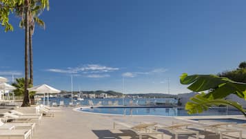A poolside area with sun loungers, a palm tree, and a view of the sea and buildings in the background.