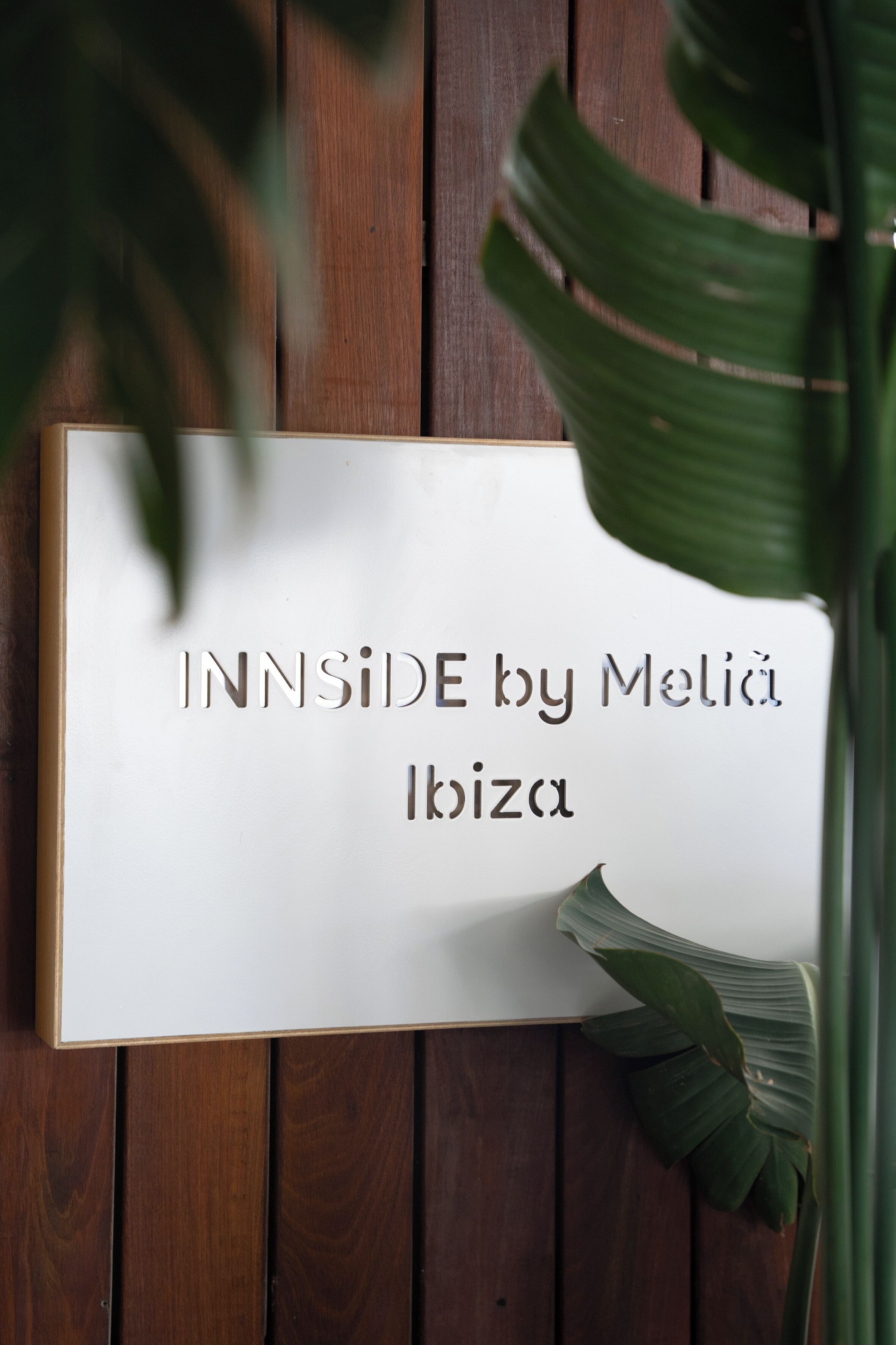 Señal con el texto 'INNSIDE by Meliá Ibiza' sobre un fondo blanco, fijada a una pared de madera con una planta parcialmente visible.