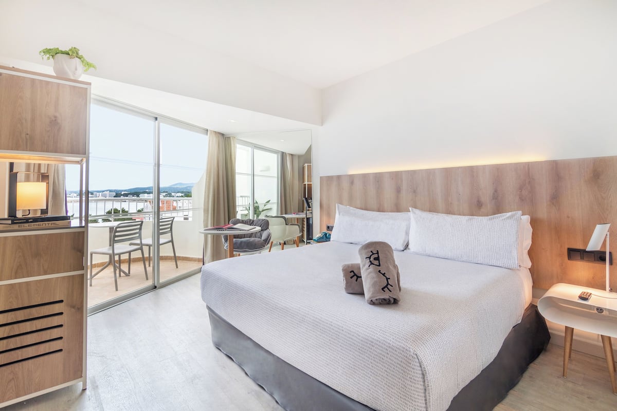 A modern hotel room with a large bed, a desk, and a balcony with a city view.